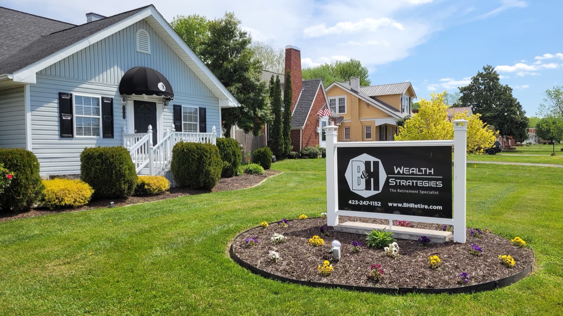 The B&H Wealth Strategies office, a pale blue house with black shutters and an awning over the front door, with the firm's sign in a flowerbed on the front lawn.